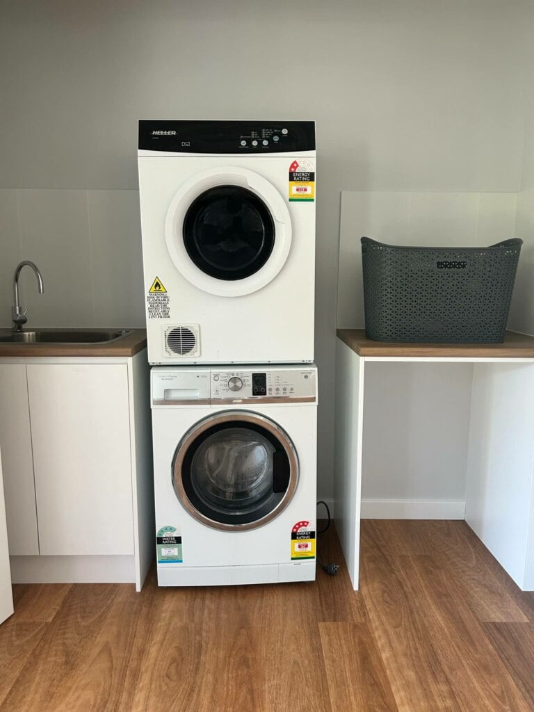 A stacked washer and dryer set, supported by independent living amenities, located in a modern laundry room with a sink and a grey laundry basket in Lethbridge Park NSW.