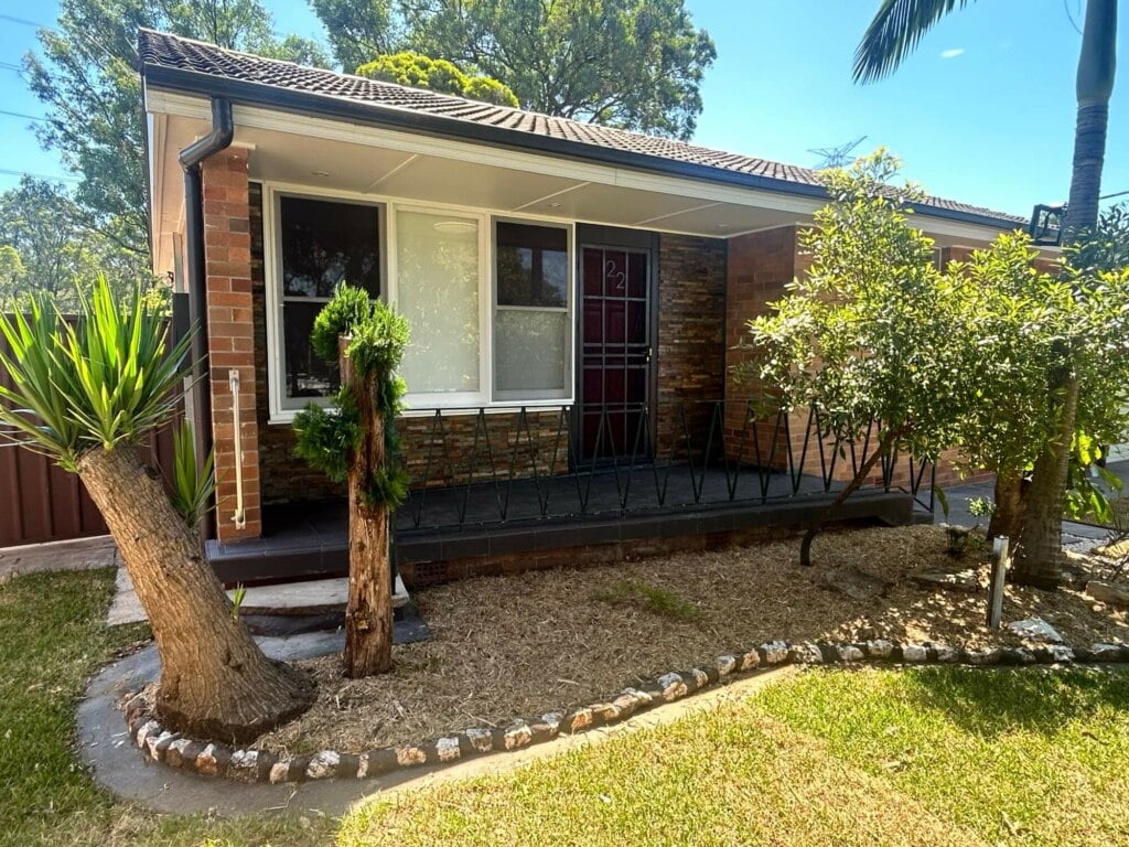 Single-story house with a brick facade and a small front porch flanked by palm trees and landscaped garden beds, offering supported independent living in Lethbridge Park NSW.