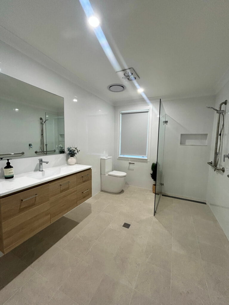 A modern bathroom in Lethbridge Park NSW, designed for supported independent living, features a large mirror, double vanity, walk-in shower, and toilet.