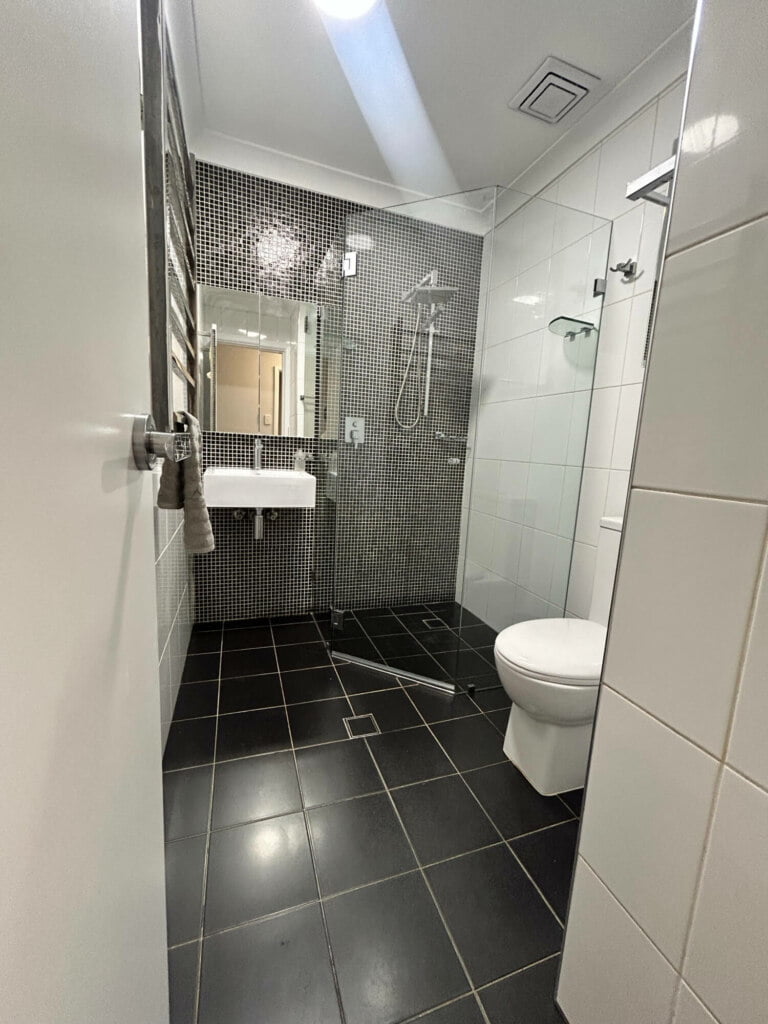 A modern bathroom with white fixtures, dark floor tiles, and a mosaic-tiled shower area, located in a supported independent living facility in Lethbridge Park NSW.