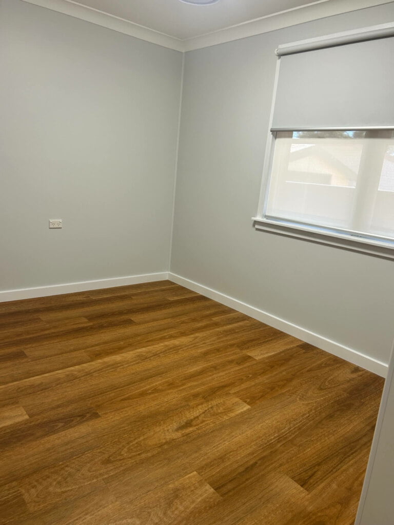 An empty room with wooden flooring and a window with a rolled-up blind, designed for supported independent living in Lethbridge Park NSW.