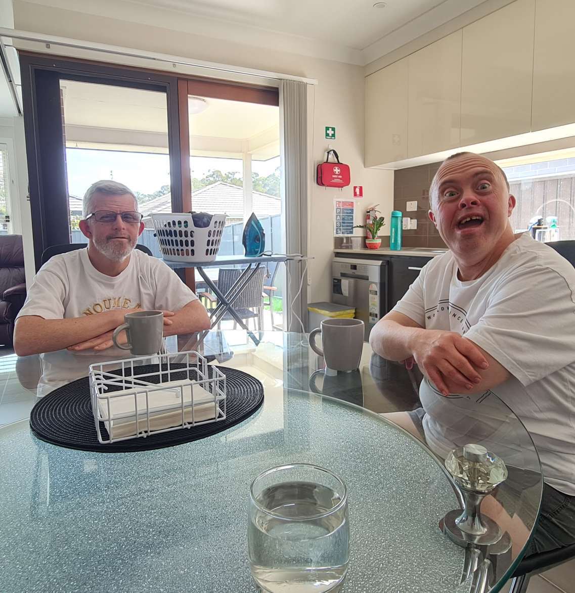 Two men seated at a kitchen table with a dishwasher basket between them; one is looking directly at the camera with a surprised expression, while the other sits calmly with his arms crossed.