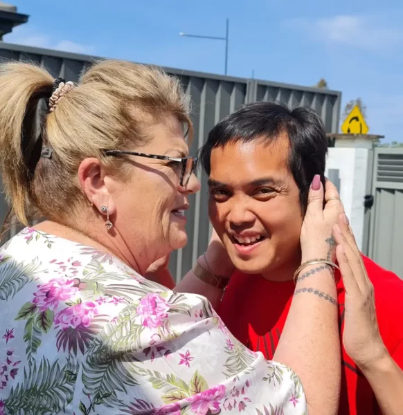 A woman in a floral shirt and sunglasses on her head is embracing a smiling man in a red shirt; both appear to be enjoying a sunny day outdoors.